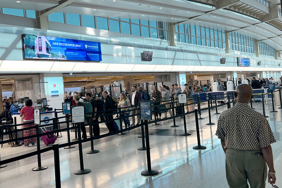 At Dulles Airport, passengers are lined up at a security checkpoint with luggage in tow. A small digital advertising display on the wall, close to the ceiling, reads 'A New Space for Hopkins Impact.'