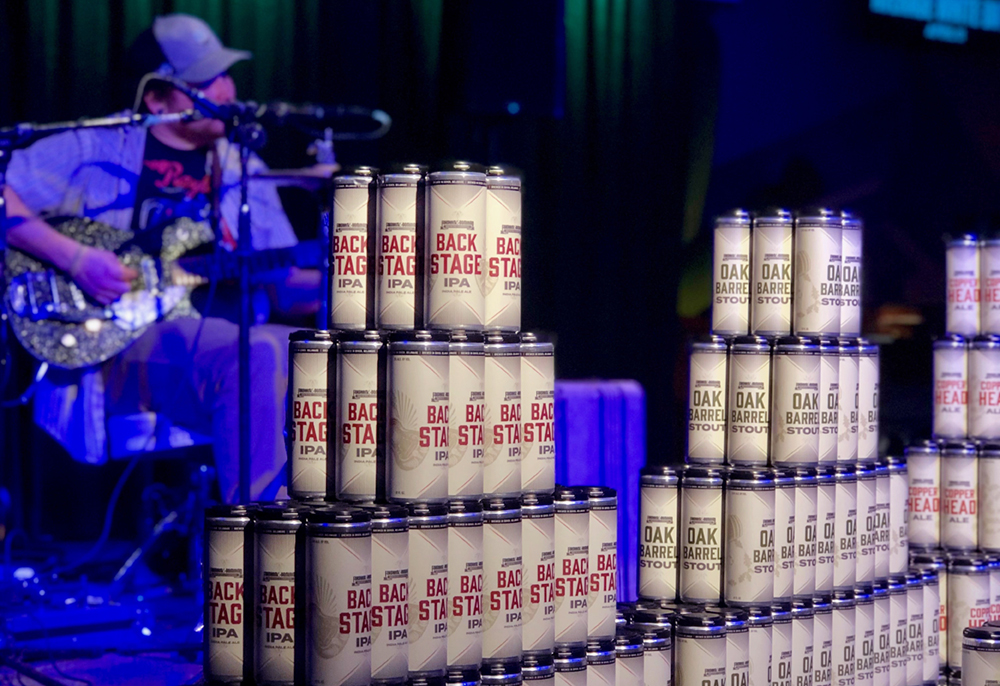 stacked beer cans with redesigned labels in front front of a live musician