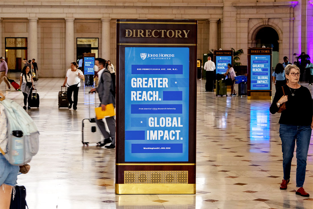 A digital poster in the main corridor of a train station reads 'Greater Reach. Global Impact.' Commuters walk past the ad with backpacks and suitcases.