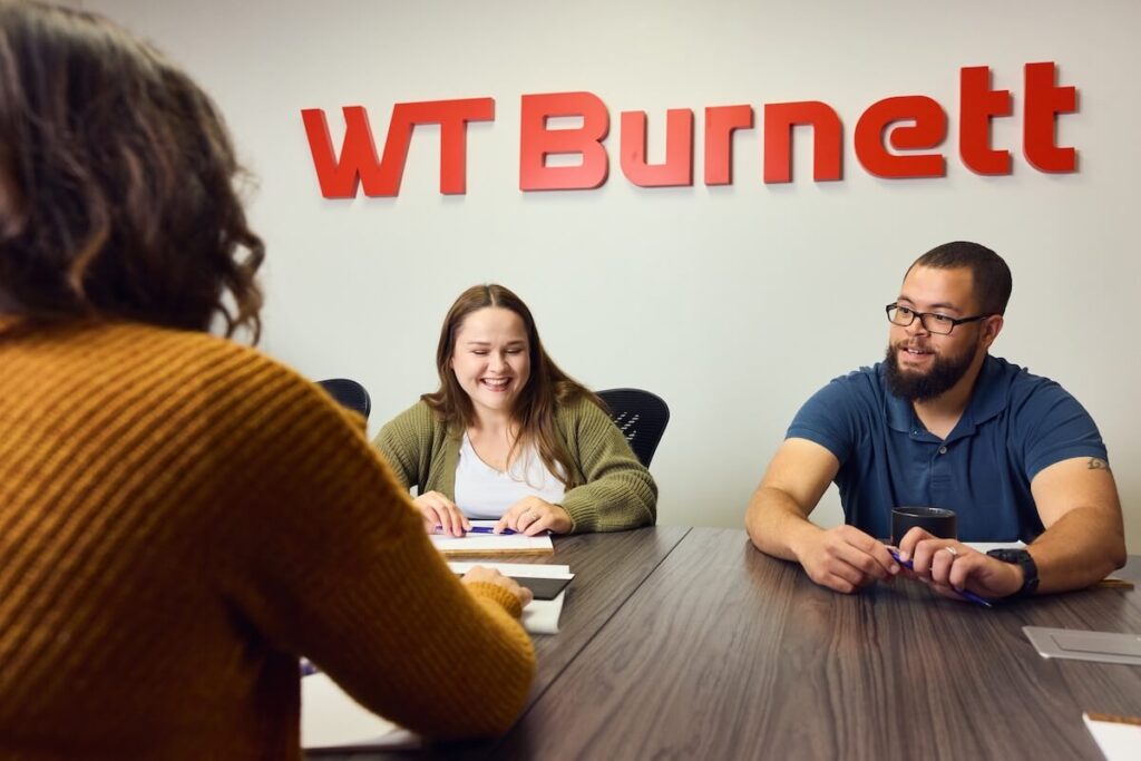 A male and female employee of WT Burnett shown sitting at a conference room table analyzing a new foam sample with new identity in the background