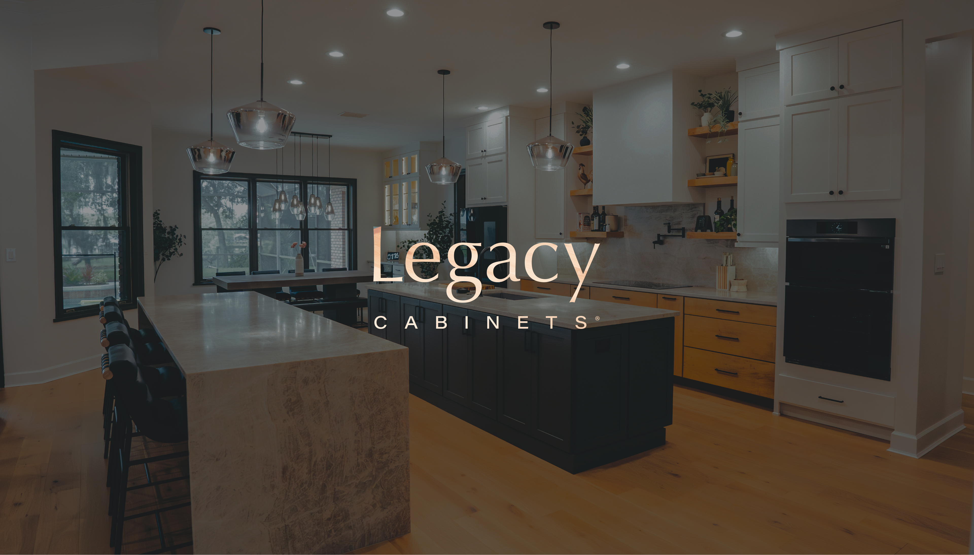 Legacy Cabinets wordmark over a modern kitchen interior featuring a navy island, light wood cabinetry, and warm pendant lighting.