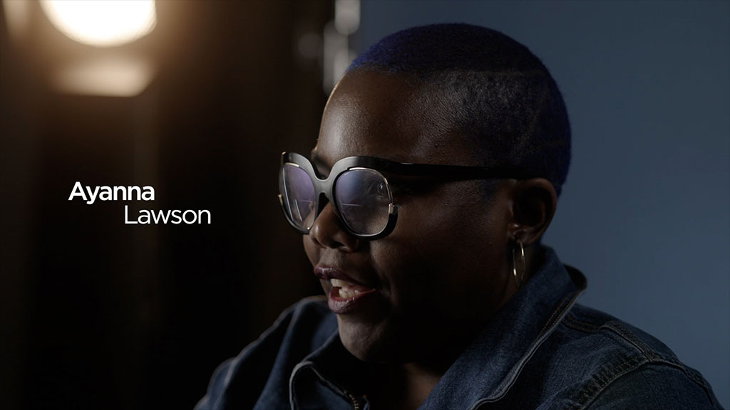 A close-up portrait of a person wearing large glasses and hoop earrings, speaking in a dimly lit studio setting. The individual is shown in profile against a dark blue background with soft lighting, while their name, “Ayanna Lawson,” appears in white text on the left side of the image. The image has a cinematic, documentary-style feel.