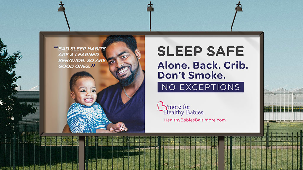 A large outdoor billboard promoting infant sleep safety, featuring a smiling father holding a young child. The billboard includes the message “Sleep Safe. Alone. Back. Crib. Don’t Smoke. No Exceptions.” alongside a quote about healthy sleep habits. The campaign branding for B’more for Healthy Babies and the website HealthyBabiesBaltimore.com appear at the bottom. The billboard is displayed in an outdoor urban setting during daylight.